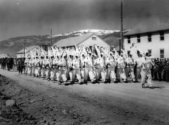 10th Mountain Division soldiers parade down a street at Camp Hale, Colorado. They are wearing their "whites" the winter camouflage uniforms and carry white skis on their right shoulder as rifles are normally carried while on parade.