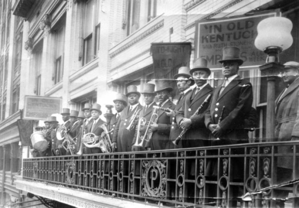 Black musicians pose on a balcony on an unidentified building in Denver, Colorado. They hold instruments that include: piccolos, clarinets, trumpets, coronets, baritones, french horns, trombones, and drums. Nearby signs read: "In Old Kentucky With Ruth Stoneho[?] All Week" and "To-night In Old Kentucky".