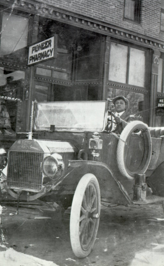 Andres "Andy" Trujillo drives his Model T Ford past the Pioneer Pharmacy in Fort Collins (Larimer County), Colorado. The young man wears a cap, coat and tie.