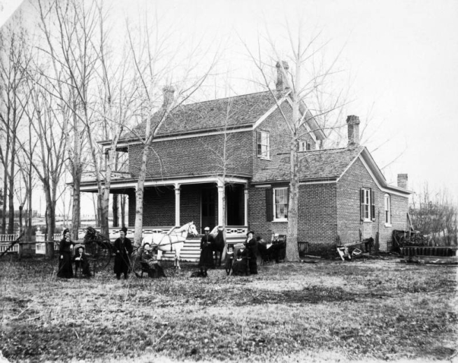 Home of Mr. Addison Nathan Baker on Grand Avenue (West Colfax Avenue) in Denver, Colorado. The farm house is a two story brick structure with a wraparound porch. The Baker family poses on the lawn, men, women, boys, and girls sit and stand, shows a dog and a horse drawn carriage.