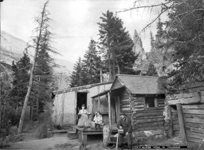 A miner and his family pose on the porch of their cabin in Colorado. The man holds a lunchbox and sits on a wheelbarrow.