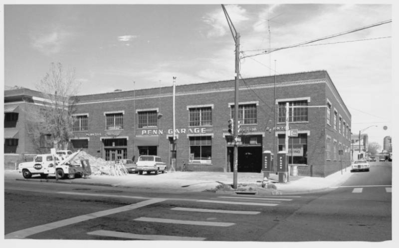 View of the two story brick Penn Garage at the corner of 13th (Thirteenth) Avenue and Pennsylvania Streets in Denver, Colorado. The car repair shop has a bay for cars, and a parking lot. A sign in a window reads "Emission Testing." The building has a flat roof and a course of dogtooth brick beneath the cornice. Phone booths are on the corner, automobiles are parked nearby, and a tow truck that reads "Penn" is in the parking lot. Shows a pile of construction debris in the parking lot.