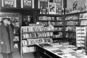Interior view of a book store in Denver, Colorado; shows books, magazines, and men identified as Detective John Wells (topcoat and fedora hat) and Sam Handler.