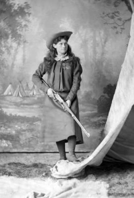 Annie Oakley, wearing hat and two medals, holding rifle in both hands, one foot propped on edge of teepee.
