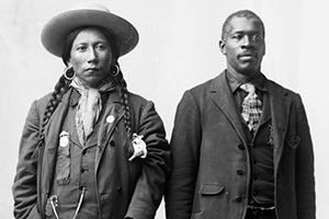 Standing portrait of a Native American Ute man, identified as Dick Charlie and an African American man, John Taylor. Both men wear vested suits; Charlie with earrings and a kerchief around his neck, Taylor with a tie.