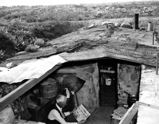 "John Baker, shown here reading his paper on the 'porch' is taking care of the home of his friend Dutch in Gopher City while Dutch is panning gold in the mountains." From the WHG Undigitized Photo files (Denver. Slums. Gopher City).