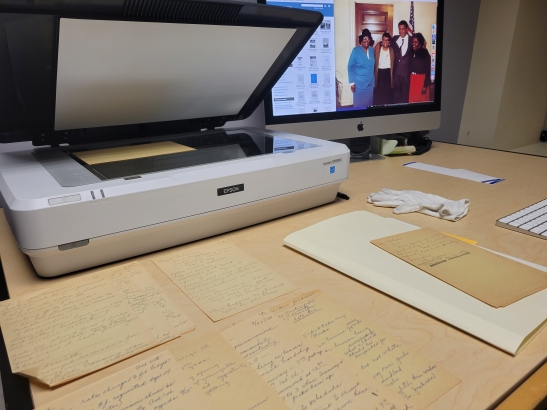 An epson scanner connected to a mac desktop, archival papers are laid out on the desk, a pair of white gloves are used to handle the archival papers.