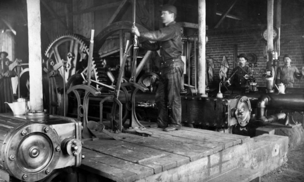 A miner stands with his hand on a large lever in the hoist house at a mine near Leadville (Lake County), Colorado. Shows large gears and heavy machinery. Workers stand near the wall.