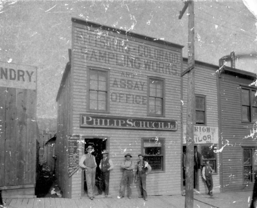Men identified as Philip Schuch Jr. and Earl Akin stand in the door of an assay office, signs read: "Pittsburg - Colorado Sampling Works and Assay Office, Philip Schuch, Jr.," Cripple Creek, Colorado. The business is housed in a false front clapboard structure.  A sign posted near the door lists prices for services. A man identified as Dean Akin stands near a window.  An unidentified man leans against the wall of the tailor shop nearby.