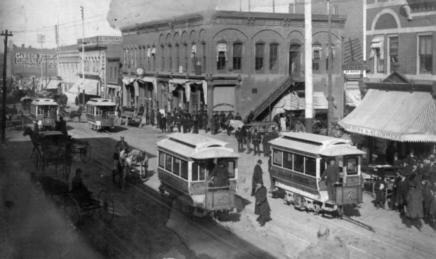 View of trolley cars on 15th (Fifteenth) Street at the intersection of Lawrence Street in downtown Denver, Colorado. Men stand beside commercial buildings; sit on horse-drawn wagons; or stand on trolley cars with numbers and lettering that read: "7," "2," "5," "6," "E. Eighteenth St.," and "Colfax Avenue." Signs painted on multi-story brick buildings read: "Garson, Kerng[ood & Co.] Clothiers, Furnisher[s]," "A. Jacobs & Co. Boy's Clothing," and "W.E. Stone Dentist" on the McClelland building.