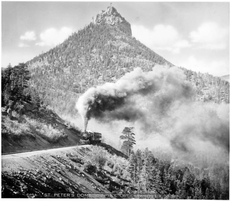 View of a smoking locomotive with passenger cars on the standard- gauge Colorado Springs & Cripple Creek District Railway; St. Peter's Dome rock formation in background with flag flying.