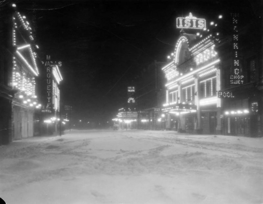 Night view of Curtis Street in Denver, Colorado; shows snow and movie theaters. Electric signs read: "Marquette," "Iris Theater," "Isis," and "The Nanking Chop Suey, Pool."