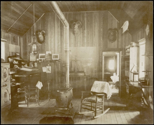 View of the interior of the Wapiti Mining Company office in Breckenridge (Summit County), Colorado. A rocking chair with a cushion sits next to a wood stove and chimney in the center of the room. A lamp hangs over a desk in the corner, next to a safe. Taxidermic heads of mountain sheep hang on the walls. Large scales sit on a table in front of map tacked to the wall. A pickaxe hangs from book shelves.