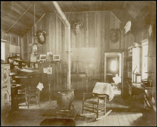 View of the interior of the Wapiti Mining Company office in Breckenridge (Summit County), Colorado. A rocking chair with a cushion sits next to a wood stove and chimney in the center of the room. A lamp hangs over a desk in the corner, next to a safe. Taxidermic heads of mountain sheep hang on the walls. Large scales sit on a table in front of map tacked to the wall. A pickaxe hangs from book shelves.