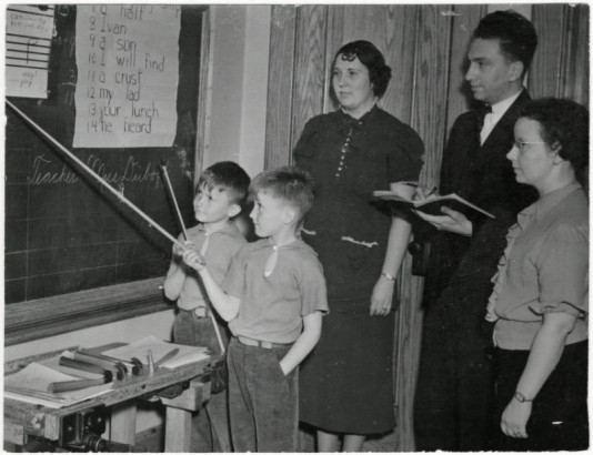 Photograph of two unidentified students, and three staff members from University Park Elementary School located in Denver, Colorado.  In the picture, two boys stand to the side of the blackboard.  They have pointers in their hands.  Two women and one man stands behind the blackboard.  The man appears to be writing something in a notebook.