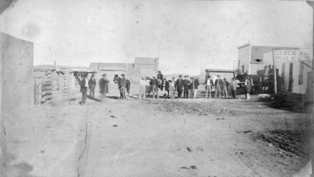 Men stand and wear hats, jackets and boots, in a dirt road near parked covered wagons and horses in Trinidad (Las Animas County), Colorado. A man poses with a horse on a lead rope. A sign supported by a timber reads: "Lunch Hou[se] Meals at all Hou[rs]" Frame, log, and an adobe building are beside the street.