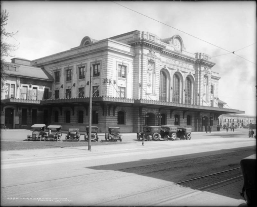 Northwestward, exterior view of the Union Station train depot in Denver, Colorado; shows automobiles parked in front of station, a boy standing on sidewalk, streetcar tracks on Wynkoop Street, and people standing in front of station at far right.