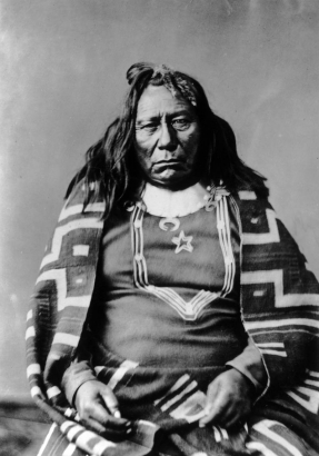 Portrait of Chief Colorow, called Colorado, Native American Ute Indian; he wears long hair with banks tied back in a fur tail and is wrapped in a woven patterned blanket.
