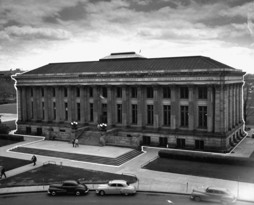 North facade of the Denver Public Library, Civic Center, Denver, Colorado. The Greek revival style Carnegie building features gray Turkey Creek Sandstone construction, fourteen fluted columns with Corinthian capitals, hipped roof and skylight. Inscription along freize reads: "Erected in the year of our Lord nineteen and nine and dedicated to the advancement of learning." Automobiles park along Colorado U. S. 40 or Colfax Avenue.