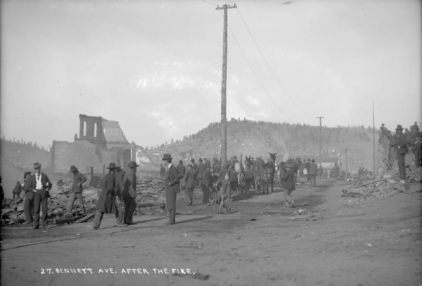 Large group of men and on-lookers view destruction, debris and remains of two-story brick wall on Bennett Avenue caused by second fire on April 29, 1896, Cripple Creek, Colorado; mule-drawn wagon loaded with wooden barrels is parked along edge of street; men are lined along railing by the elevated portion of Bennett Avenue; Tutt-Penrose building and other commercial business buildings untouched by fire.