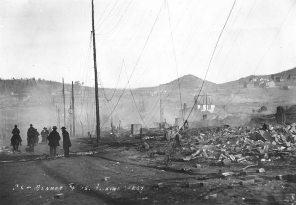Destruction west on Bennett Street caused by second fire on April 29, 1986, Cripple Creek, Colorado; groups of men and two men on horseback in street survey rubble and remains of once thriving commerical business district; shows utility poles with fallen wires, chimneystack, remains of two-story brick building, Mt. Pisgah is on horizon and untouched  residences are on hillside in background.