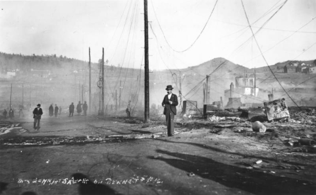 View of destruction west on Bennett Avenue, Cripple Creek, Colorado, caused by second fire on April 29, 1896; groups of men and solitary men survey remaining debris; smoke still rises from ashes and utility poles with fallen wires border street; Mt. Pisgah and undamaged buildings are in background.