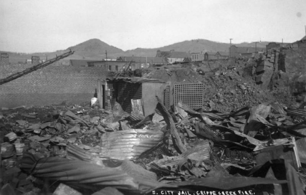 Remains of city jail demolished by first fire, April 25, 1896, Cripple Creek, Colorado; debris includes twisted corrugated tin metal, cage doors and cell block; one brick wall remains; buildings in commercial business district are in the background.