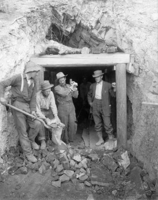Men pose by the Lady Belle mine entrance south of Eagle, Eagle County, Colorado; one man shovels ore into a canvas bag.