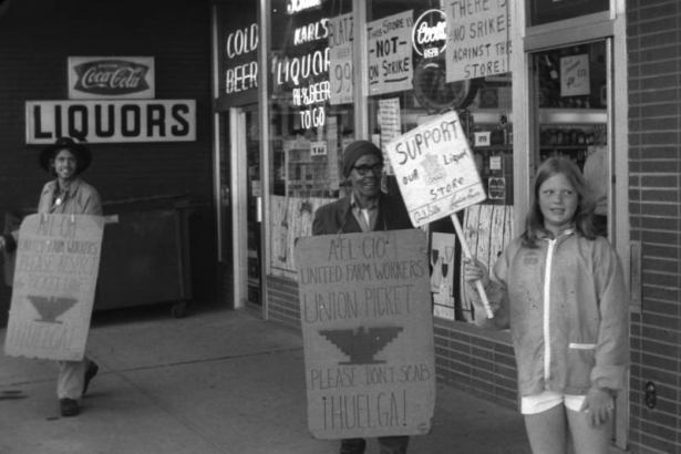 Jesús Valderrama pickets Karl's Liquors for the sale of Gallo Wine at West Colfax in Denver, Colorado. He holds a sign that reads: "AFL-CIO United Farm Workers Union Picket. Please Don't Scab. Huelga."  A young girl, the daughter of the owner of Karl's Liquors, stands beside him with a sign that reads: "Support Our Liquor Store."  A second protester is in the distance. Signs in the window of the store read: "This Store Is Not On Strike" and " There Is No Strike Against This Store."