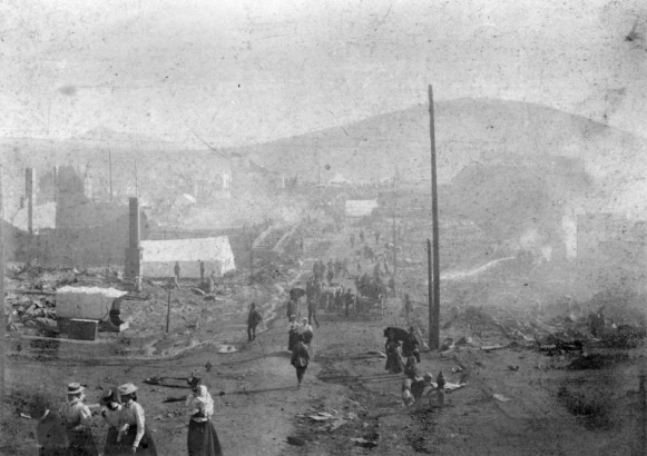 Aftermath of destruction caused by April, 1896 fire, Cripple Creek, Colorado; men, women and children stroll down street that has debris  and rubble left from fire; foreground includes woman holding baby, boy drinking from fire hydrant, canvas tents amidst rubble, and stream of water  sprayed on rubble.