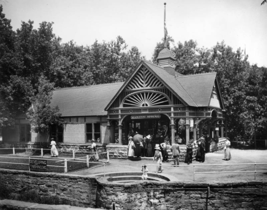 Tourists come and go from the Soda Spring Pavilion, Manitou Springs, El Paso County, Colorado. The open air pavilion features decorative woodwork with sunburts, rusticated stone foundation, roof cresting, four sided dome with flag pole and fish scale shingles. Signs read: "Manitou Springs," and "Developing and Finishing Films and Supplies."