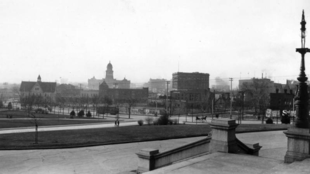 View looking northwest towards downtown from State Capitol steps, Denver, Colorado, includes cable street cars, carriages, Engine Company No. 1's victorian firehouse across the intersection of Broadway and Colfax and the Arapahoe County Courthouse in distant haze. Signs on billboards and foreground businesses read: "Palace Stables," "Plymouth Hotel," "$25.00 Rio Grande," "Denver Clothes Pressing Co., $3.00 for 3 Months," "Automobiles, Repairs, Storage."