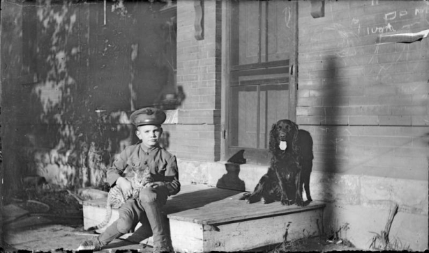 Outdoor portrait of a boy in Denver, Colorado, wearing a World War One or Western Union Telegraph uniform. He holds a tabby cat; an Irish Setter is by a brick wall with graffiti.