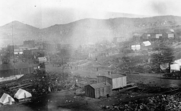 View northwest towards Bennett Avenue after destruction from second fire of April 29, 1869, Cripple Creek, Colorado; shows canvas tents on edge of debris, wood frame buildings under construction, people walking down street, crowd gathered at distant corner, and Mt. Pisgah on horizon.