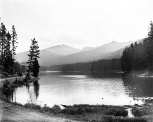 A man sits in a touring automobile on Cody Road, along Sylvan Lake, Yellowstone National Park, Wyoming; Top Notch Peak shows in distance.