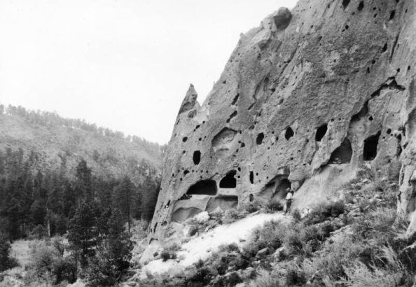A Native American man stands next to a tuff cliffside with eroded holes enlarged by ancestral Pueblo Indians for dwellings and ceremonial spaces, Pajarito Plateau, New Mexico; became Bandelier National Monument in 1916.