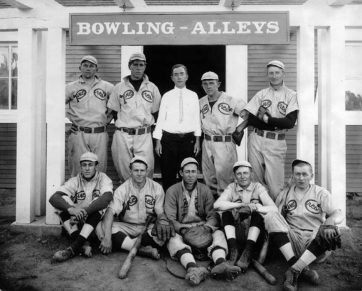 The Pine Crest Baseball team poses in front of the Palmer Lake Bowling Alleys, El Paso County, Colorado. They wear uniforms (caps, shoes with cleats, baseball gloves.) The catcher with his mitt sits front and center; players next to him have wooden bats. Possibly the coach or owner of the bowling alley stands in center back row. He wears a white shirt and striped tie.