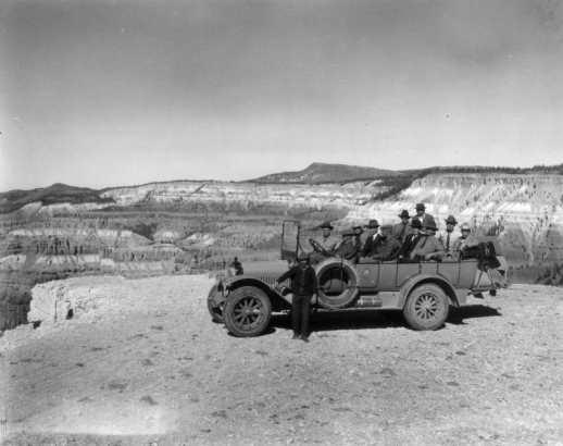 Group shot of possibly President Harding and cabinet members on their 1923 tour of Yellowstone National Park, Wyoming. The driver stands beside the long touring automobile filled with men, parked on at a lookout over eroded canyon cliffs. Emblem on door has a bear and reads: Y. P. T. Co. No. 143.