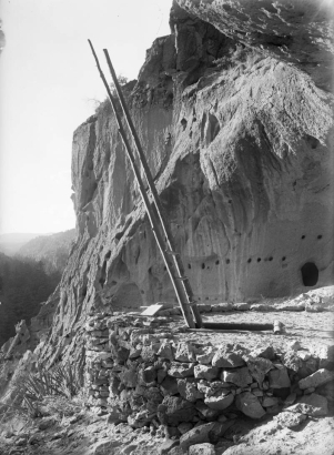 A timber ladder provides access into a kiva structure under volcanic tuff cliffs, Frijoles Canyon, Bandelier National Monument, New Mexico. The reconstructed kiva is built up with rock and has a rectangular entrance framed with timber. A natural cave enlarged by ancestral Pueblo Indians shows in background.