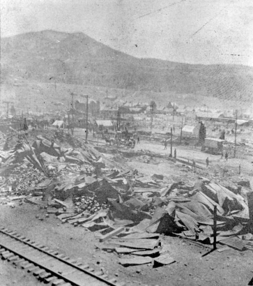 Rubble and destruction caused by second fire of April 29, 1896, Cripple Creek, Colorado; piles of twisted sheet metal, canvas tents amidst rubble, railroad tracks in foreground; view across fire devastated area toward distant view of remaining buildings; horse-drawn wagons and carriages on street.