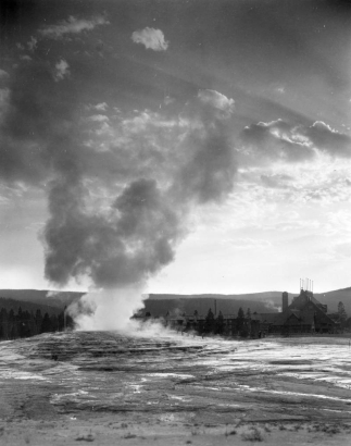 View southwest over Old Faithful Geyser to Old Faithful Inn, with steep gabled roof and a large rooftop observation platform with six flagpoles, Upper Geyser Basin, Yellowstone National Park, Wyoming. Tourists climb the terraces around the steaming geyser.