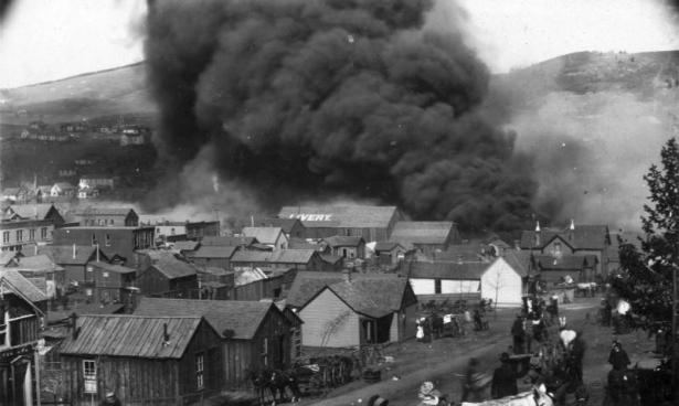 Heavy, black smoke and flames approach El Paso Livery Stable, Bennett Avenue between 1st (First) and "A" Streets, Cripple Creek, Colorado, during second fire, April 29, 1896; scene includes wood frame residences, two-story flat-roof commercial buildings and horse-drawn wagons  loaded with personal possessions ; town residents watch the approaching smoke.