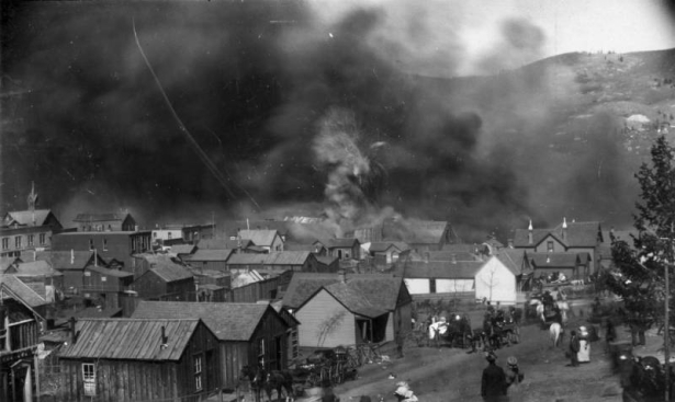 View towards El Paso Livery Stable, Bennett Avenue between 1st (First) & "A" Streets, as fire causes stored dynamite to explode during second fire on April 29, 1896, Cripple Creek, Colorado; actual blast pictured with roof debris in air, black smoke covering view of town, numerous wood frame residences and buildings in path of fire, and horse- drawn wagons packed with personal belongings; families watch approaching smoke and flames.