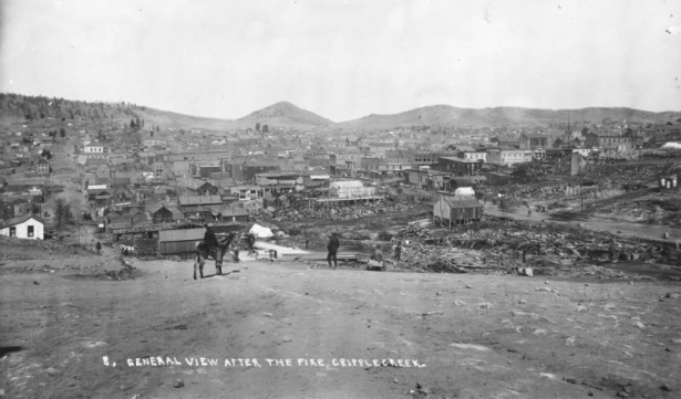 View west from Myers Avenue (probably) towards Bennett Avenue with Mt. Pisgah on horizon after first fire of April 25, 1896, Cripple Creek, Colorado; scene includes debris and  remains from fire, construction  of wood frame buildings and canvas tents and a distant view of numerous remaining wood frame and brick commercial buildings in business district; signs on buildings include: "Metropolitan House," "El Paso Livery," "Drugs," and "One price to All-King Cash-Our Motto-Weinberg Shoes & Clothing Co."