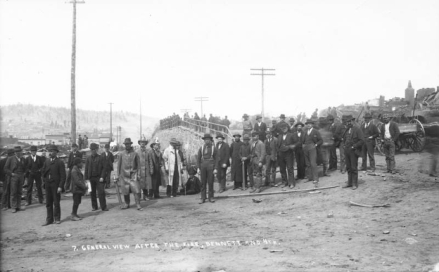 A large group of men, many firemen, survey the destruction caused by the first fire, April 25, 1896, near elevated portion of Bennett Avenue at 4th (Fourth) Street, Cripple Creek, Colorado; several firefighters wear helmets, raincoats and boots; two hold nozzles used to direct water spray; also includes horse-drawn wagon, crowd of people standing on elevated portion of Bennett Avenue and remains of commercial business district.