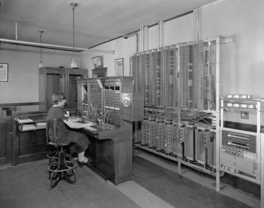 An operator works at a switchboard at the Mountain States telephone exchange building in Limon (Lincoln County), Colorado. Shows telephone equipment and telephone booths on the far end of the room. The woman wears a headset and polka-dot dress.