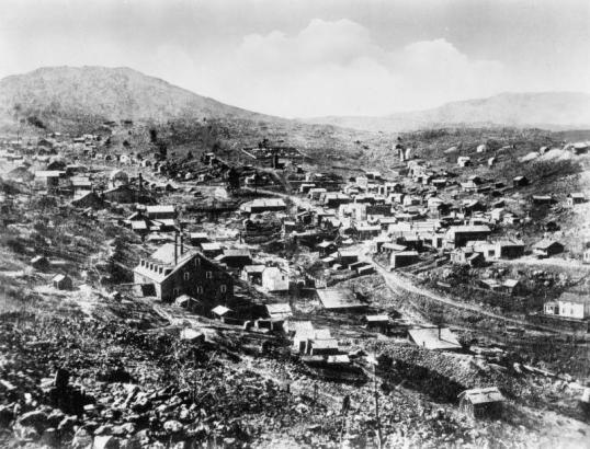 View of Nevadaville, Gilpin County, Colorado, shows dwellings, commercial buildings, The Christ Church with its square tower and steeple, Pozo Mill by Nevada Creek, mining facilities, ore shed and a fence enclosure on King's Flat with drying laundry.