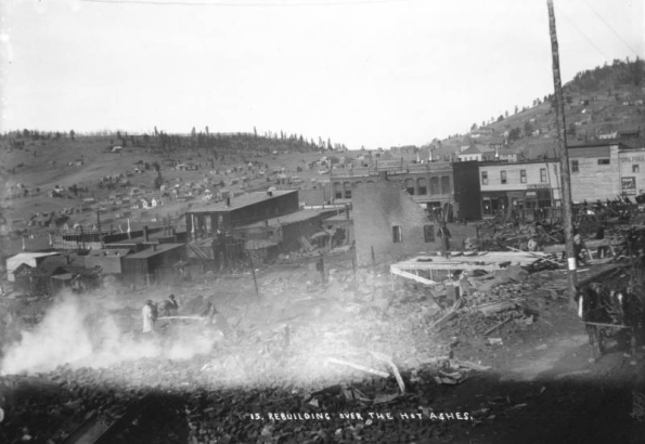 View of smouldering remains from first fire on April 25th, 1896, Cripple Creek, Colorado; three men look over debris and one man sprays water over building remains; flat roof brick and wooden frame false front buildings and the Tutt & Penrose building are in the commercial district on  Bennett Avenue; wood frame residences are on hillside in the background.
