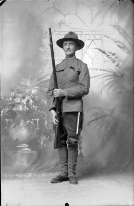 A man wears a World War I soldier's uniform and stands at attention in a studio portrait. The soldier holds a rifle and wears jodhpur pants, boots with gaiters, a long buttoned jacket and a felt hat.