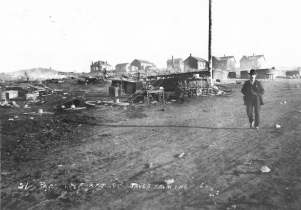 Destruction on First Street caused by second fire, April 29, 1896, Cripple Creek, Colorado; remains include stone or cement block foundations, clustered groups of high-backed chairs, tables, stool, wood commode and wagon wheels; man walks down dirt street; one-story gabled and hipped roof residences are in background.